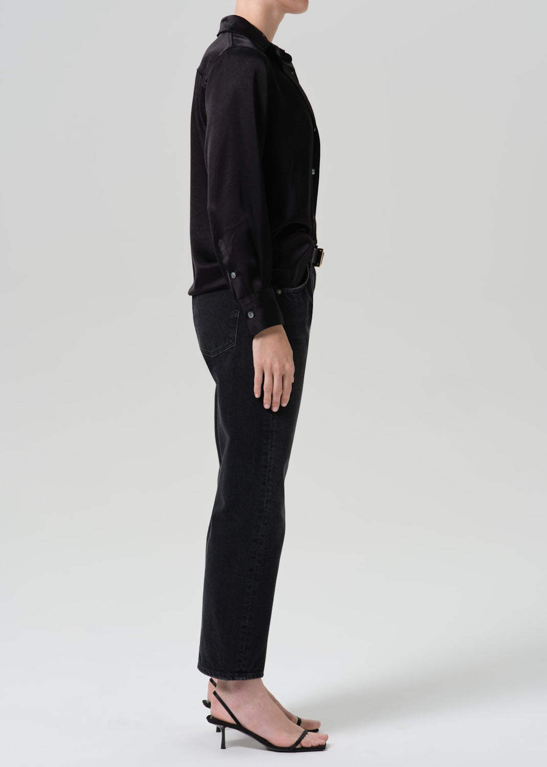 A model shown in profile wearing a black long-sleeve button-up shirt tucked into black straight-leg jeans, paired with black strappy high-heeled sandals, against a grey background.
