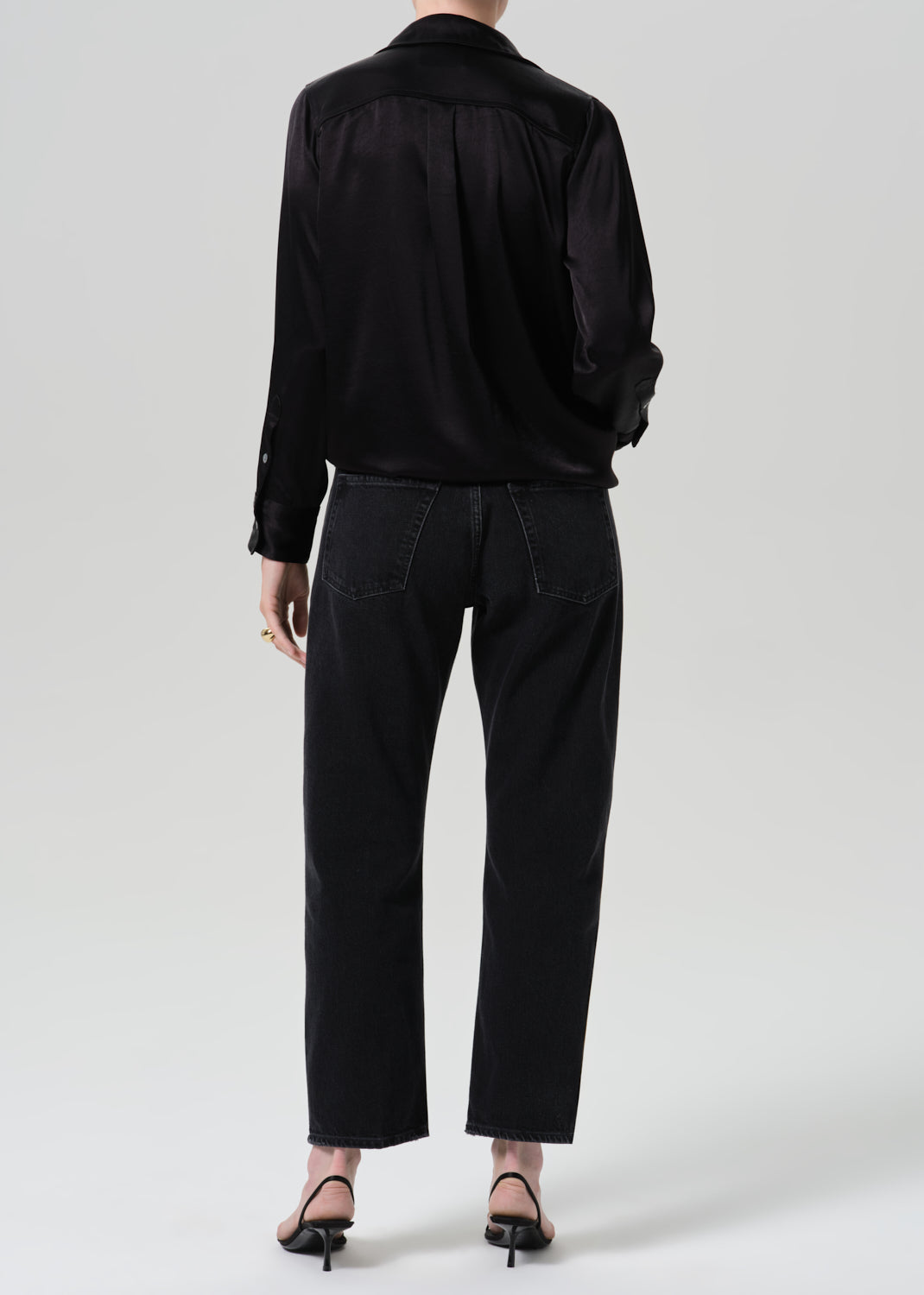 A model shown from the back wearing a black satin long-sleeve shirt and black wide-leg cropped jeans, paired with black pointed-toe heels, against a grey background.