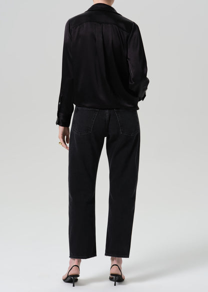 A model shown from the back wearing a black satin long-sleeve shirt and black wide-leg cropped jeans, paired with black pointed-toe heels, against a grey background.