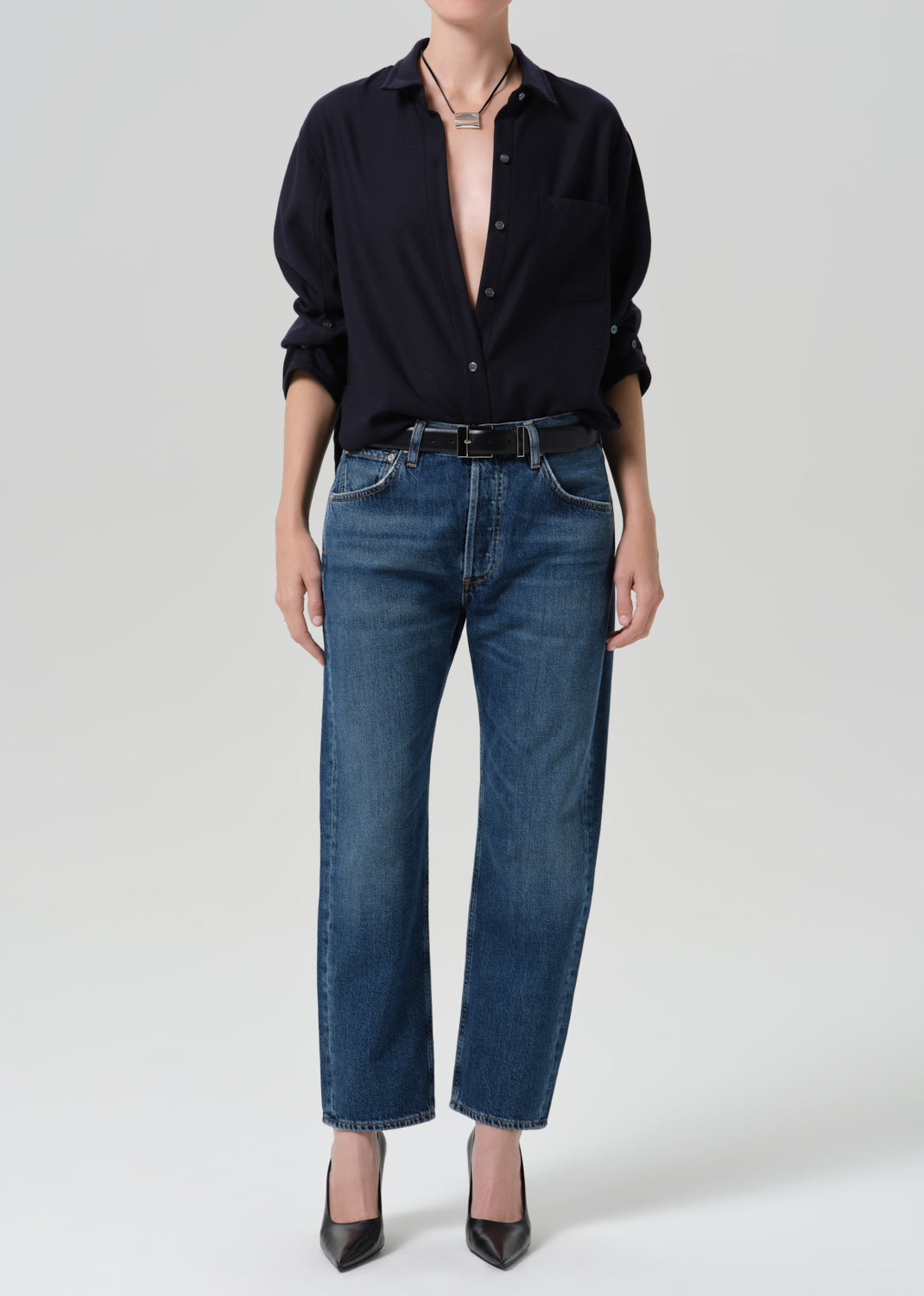 A model wearing a dark navy button-down shirt, blue straight-leg jeans, a black belt, and black pointed-toe heels, photographed against a grey background.