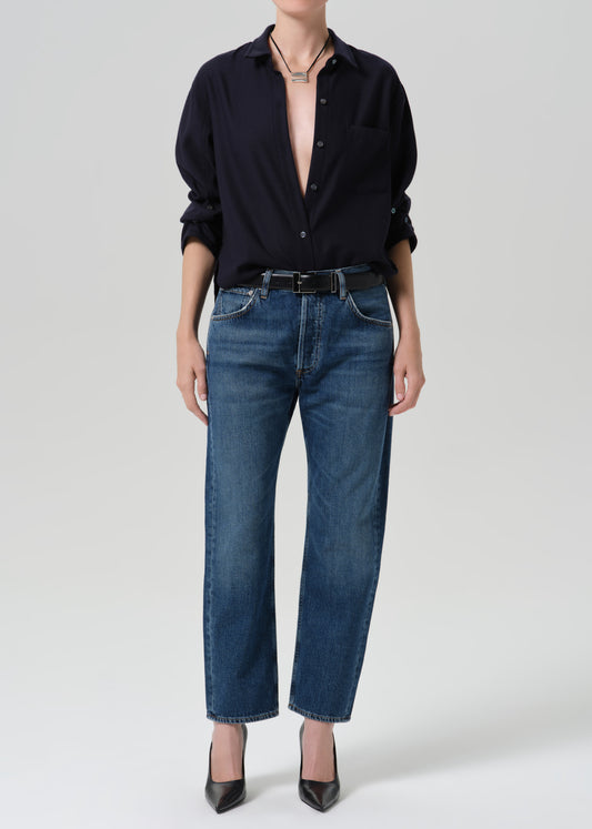 A model wearing a dark navy button-down shirt, blue straight-leg jeans, a black belt, and black pointed-toe heels, photographed against a grey background.