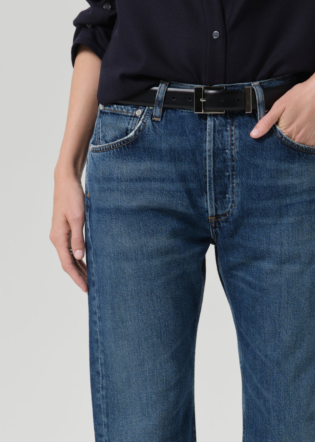 A close-up of a person wearing blue denim jeans with a black belt and a dark navy button-up shirt, shown from the chest down against a grey background.