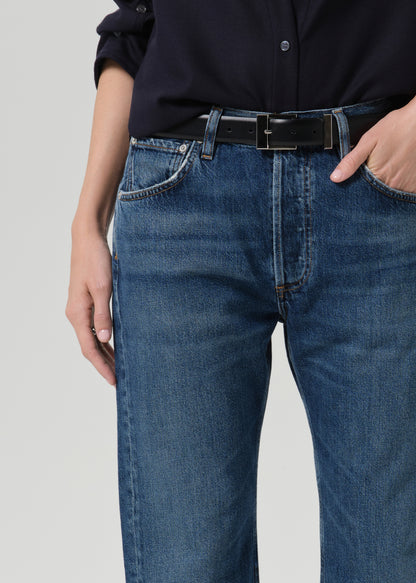 A close-up of a person wearing blue denim jeans with a black belt and a dark navy button-up shirt, shown from the chest down against a grey background.