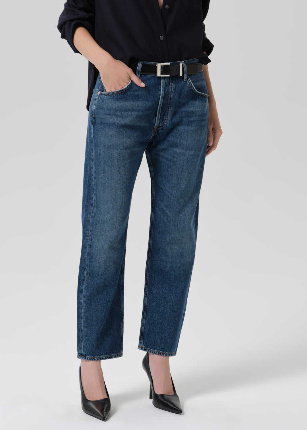A model wearing a pair of dark blue straight-leg jeans with a black belt, paired with a black shirt and black pointed-toe heels, shown from the waist down on a grey background.