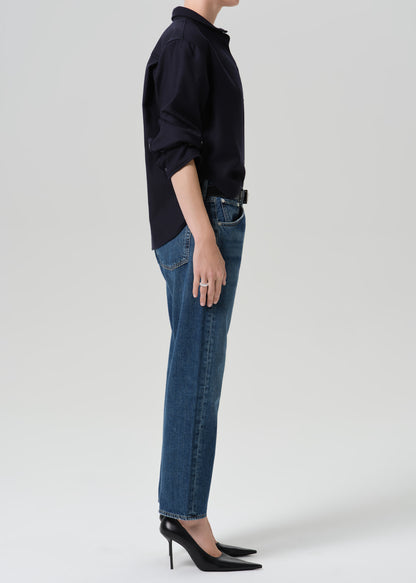 A side-profile studio shot of a model wearing a dark navy long-sleeve button-up shirt, blue straight-leg jeans, and black pointed-toe stiletto heels against a grey background.