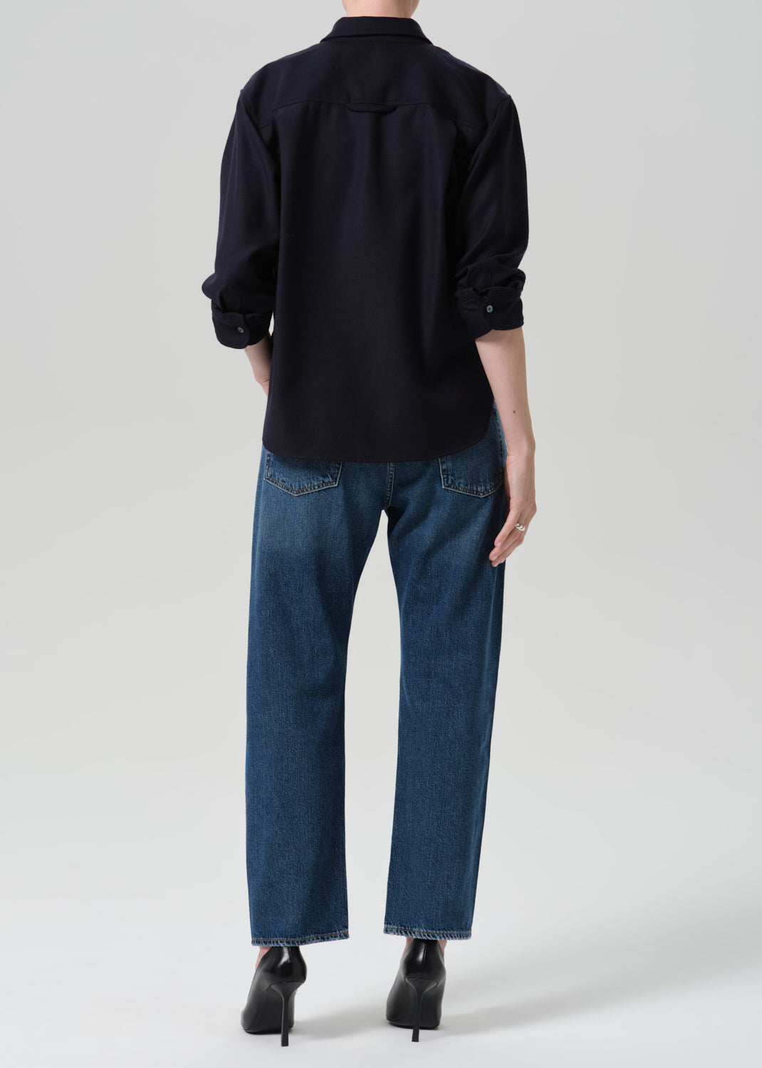 A model shown from the back wearing a dark navy long-sleeve button-up shirt, blue jeans, and black high-heeled boots against a grey background.
