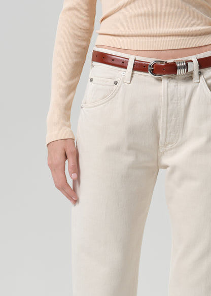 A close-up of a person wearing off-white or cream-colored jeans with a brown leather belt and a beige ribbed long-sleeve top, photographed against a grey background.