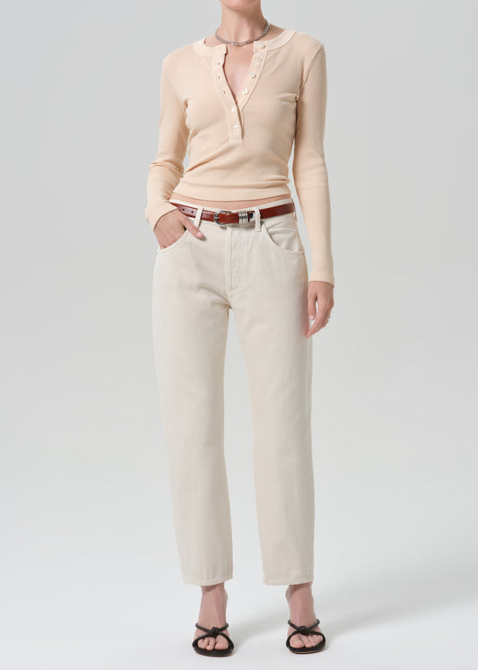 A model wearing a light beige long-sleeve knit top with a split neckline, paired with off-white cropped jeans, a brown belt, and black sandals, photographed against a grey background.