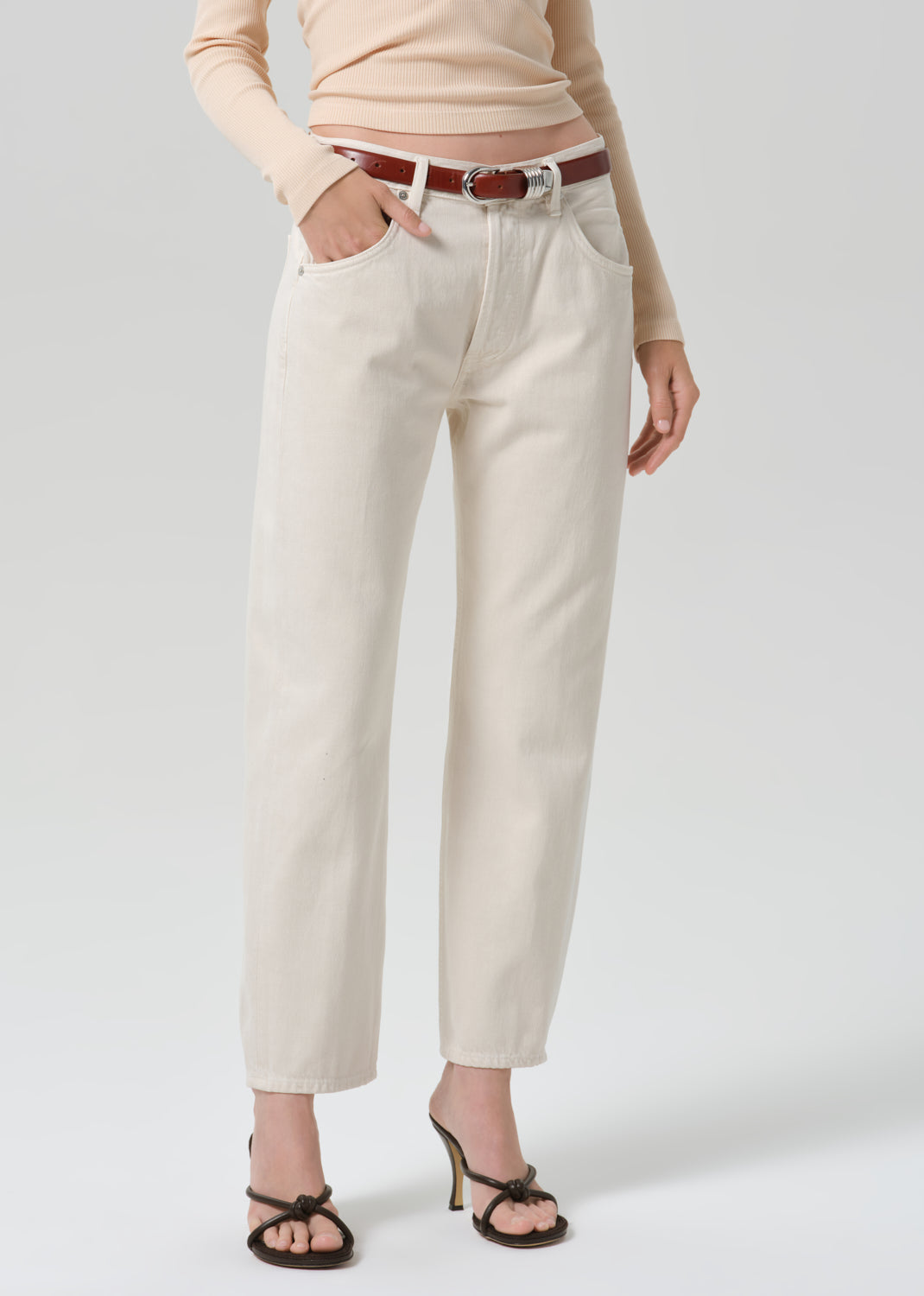 A model wearing off-white cropped jeans with a brown belt, a beige long-sleeve top, and black strappy heels, shown from the waist down against a grey background.