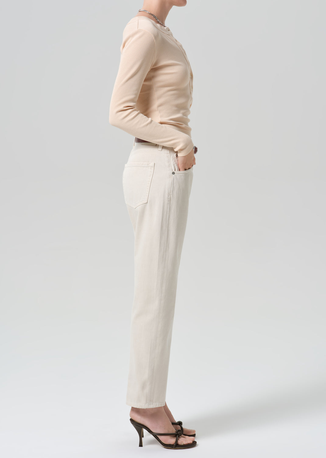 A model shown from the side wearing a beige long-sleeve top, off-white straight-leg trousers, and black strappy high-heeled sandals against a grey background.