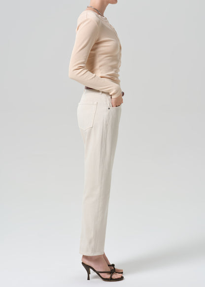 A model shown from the side wearing a beige long-sleeve top, off-white straight-leg trousers, and black strappy high-heeled sandals against a grey background.