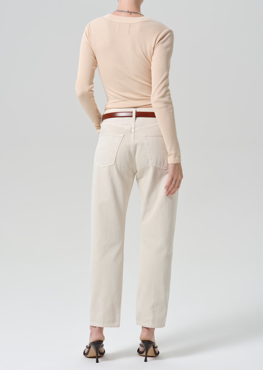 A model shown from the back wearing a light peach long-sleeve top, off-white straight-leg jeans with a brown leather belt, and black heeled sandals against a grey background.