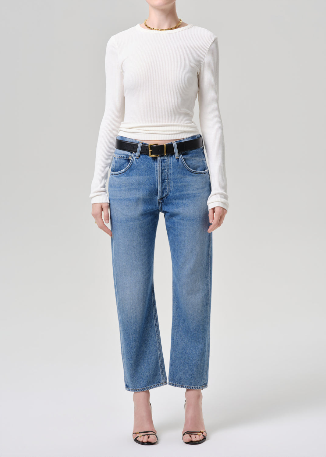 A model wearing a white long-sleeve top, blue cropped wide-leg jeans with a black belt, and black sandals, standing against a grey background.