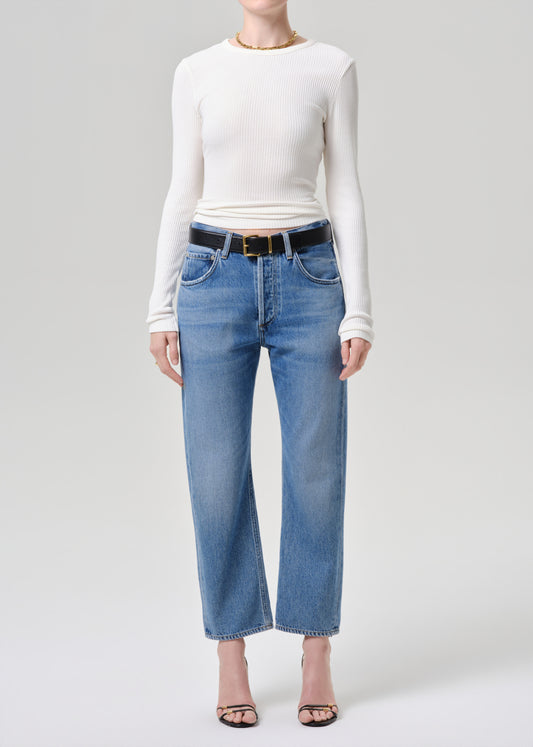 A model wearing a white long-sleeve top, blue cropped wide-leg jeans with a black belt, and black sandals, standing against a grey background.