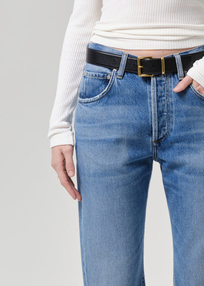 A close-up of a person wearing light blue denim jeans, a black leather belt with a gold buckle, and a white ribbed long-sleeve top against a grey background.