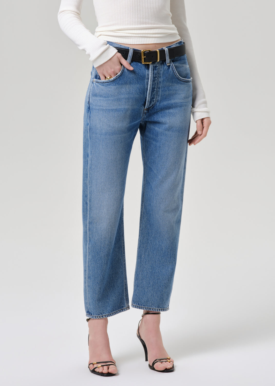 A model wearing high-waisted, wide-leg blue denim jeans with a black belt and gold buckle, styled with a white ribbed long-sleeve top and black strappy heels against a grey background.