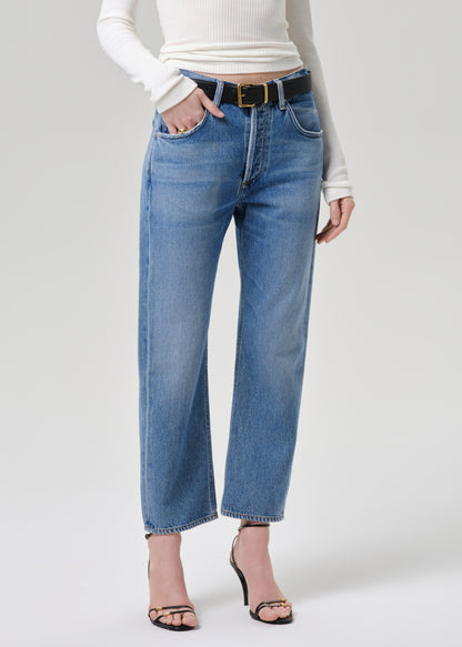 A model wearing high-waisted, wide-leg blue denim jeans with a black belt and gold buckle, styled with a white ribbed long-sleeve top and black strappy heels against a grey background.