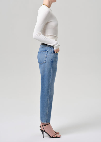 A side-profile studio shot of a model wearing light-wash blue jeans, a white ribbed long-sleeve top, and black strappy high-heeled sandals against a grey background.