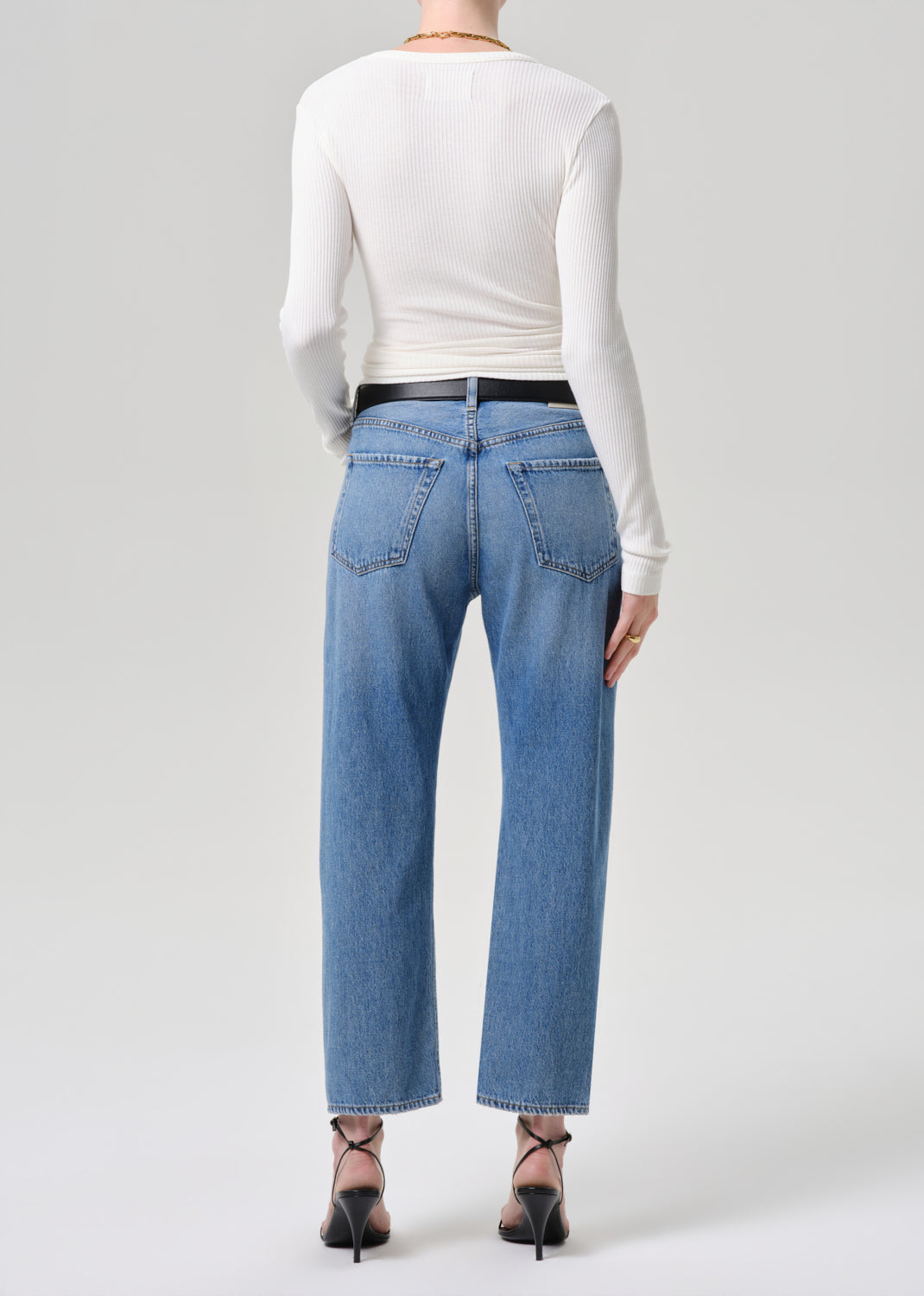 A model shown from the back wearing light-wash blue straight-leg jeans, a white long-sleeve top, and black high heels against a grey background.