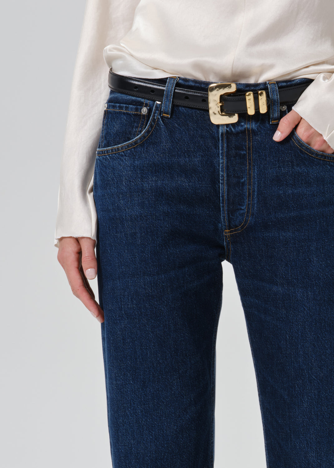 Close-up of a person wearing dark blue denim jeans with a black belt featuring a prominent gold-tone decorative buckle, paired with a cream blouse.