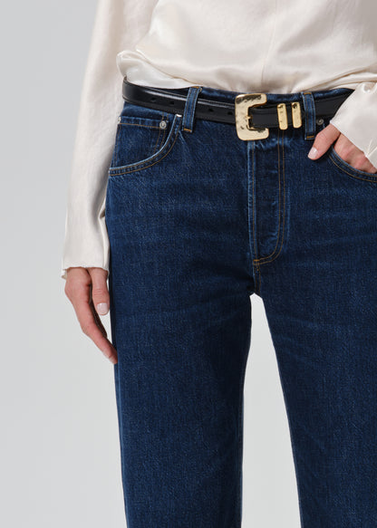 Close-up of a person wearing dark blue denim jeans with a black belt featuring a prominent gold-tone decorative buckle, paired with a cream blouse.