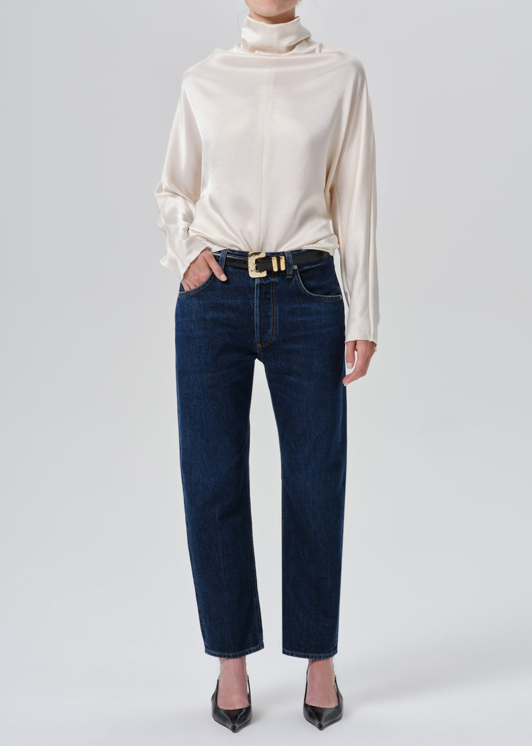 A model wearing a cream satin long-sleeve turtleneck top, dark blue straight-leg jeans with a gold-tone belt, and black pointed-toe heels, photographed against a grey background.