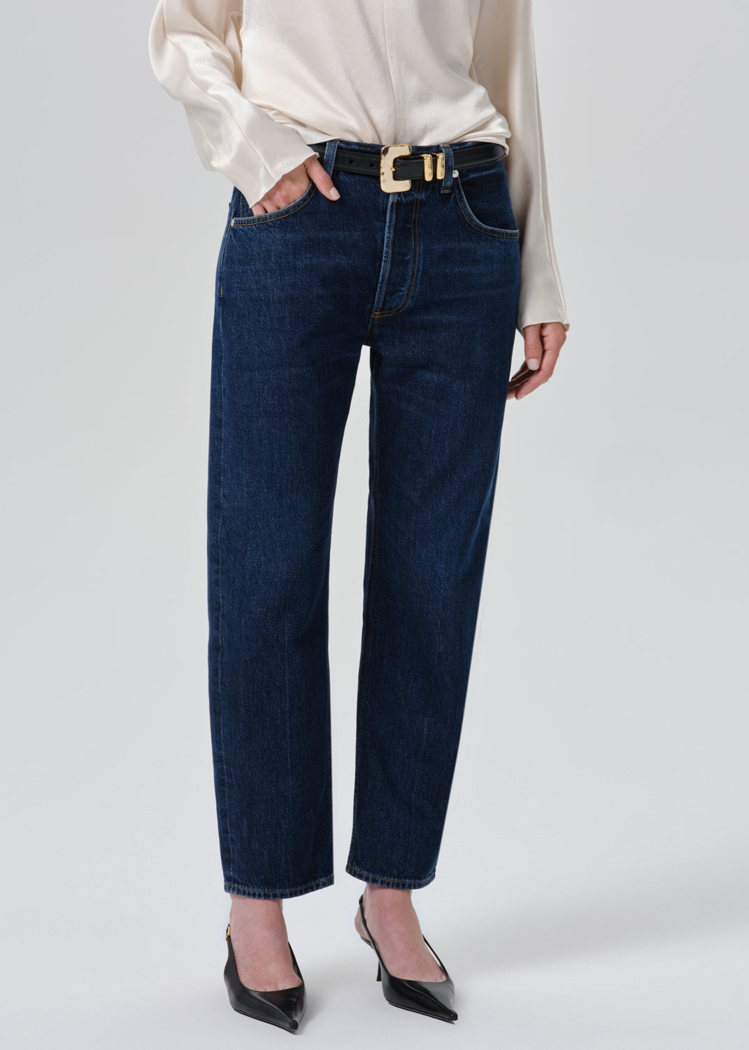 A model wearing dark blue straight-leg jeans with a gold-tone buckle belt, styled with a cream satin blouse and black pointed-toe heels against a grey background.