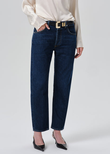 A model wearing dark blue straight-leg jeans with a gold-tone buckle belt, styled with a cream satin blouse and black pointed-toe heels against a grey background.