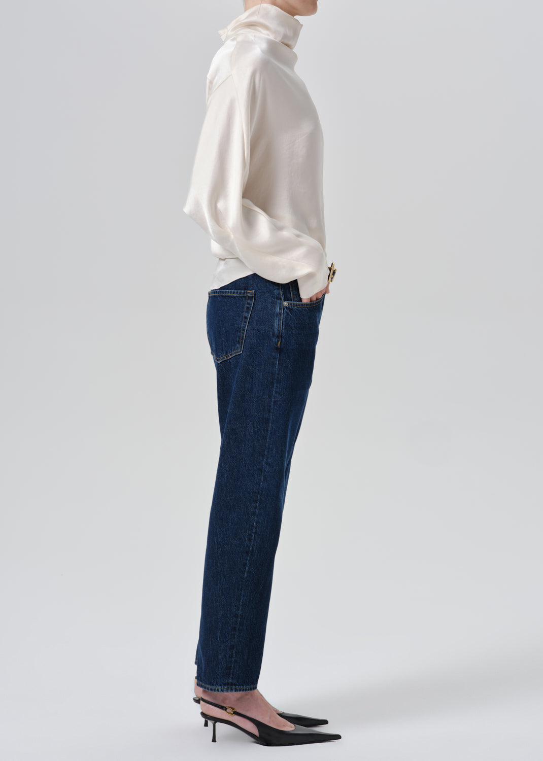 A side-profile studio shot of a model wearing dark blue straight-leg jeans, a cream satin blouse, and black pointed-toe slingback heels against a grey background.