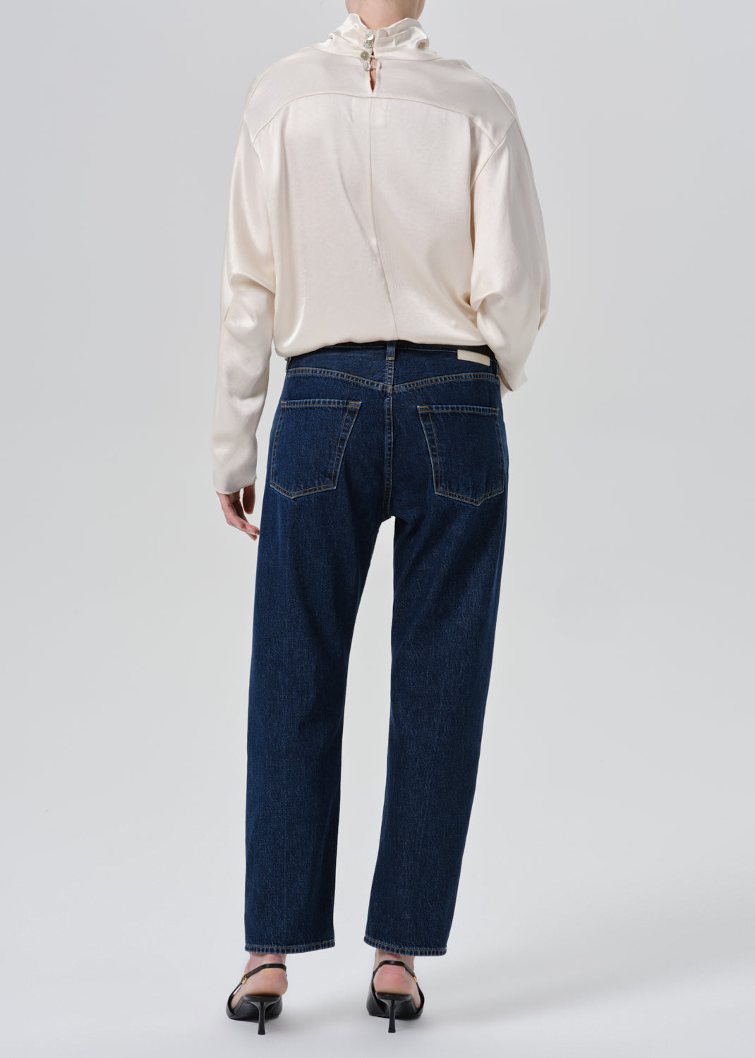 A model shown from the back wearing dark blue straight-leg jeans, a cream satin long-sleeve blouse, and black heels against a grey background.