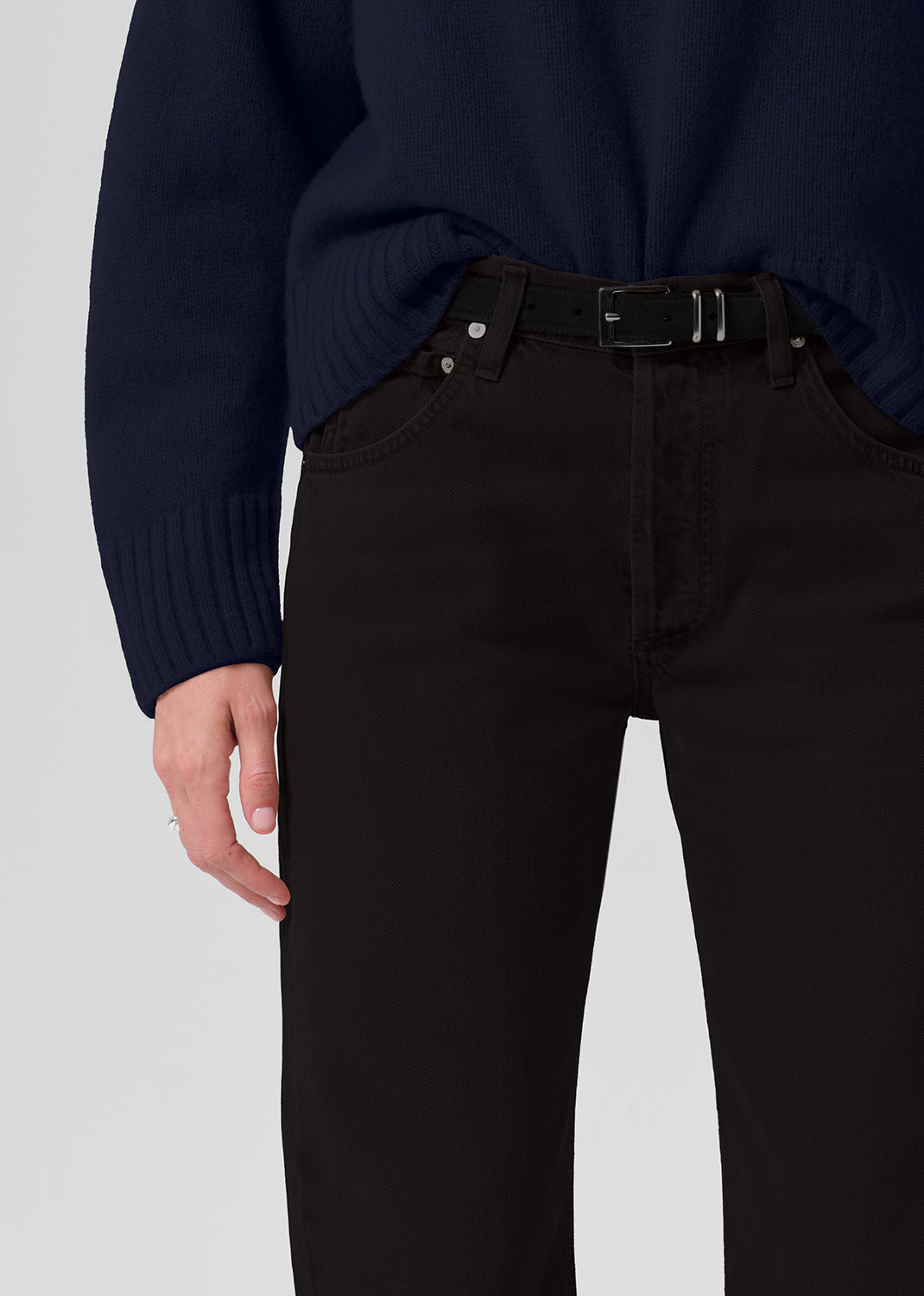 Close-up of a person wearing black jeans with a black belt and a dark navy sweater, shown from the chest down against a grey background.