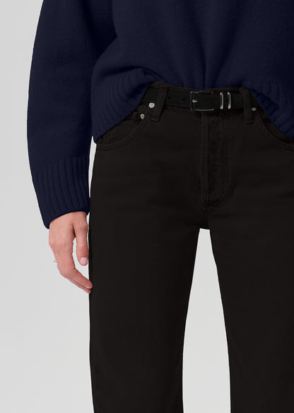 Close-up of a person wearing black jeans with a black belt and a dark navy sweater, shown from the chest down against a grey background.