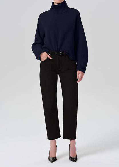 A model wearing a navy blue ribbed turtleneck sweater, black cropped trousers, and black pointed-toe heels, standing against a grey background.