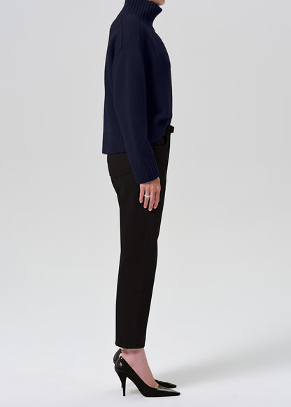 A side-profile studio shot of a model wearing a navy blue ribbed turtleneck sweater, black cropped trousers, and black pointed-toe heels against a grey background.