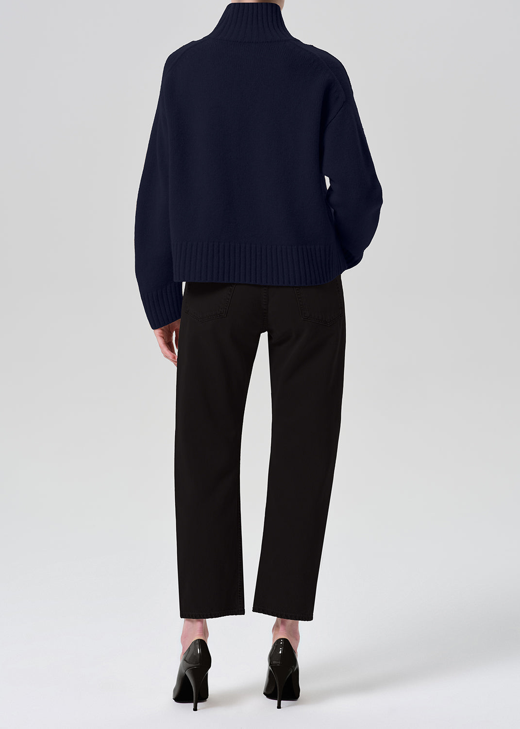 A model shown from the back wearing a navy blue ribbed turtleneck sweater, black cropped trousers, and black high heels against a grey background.