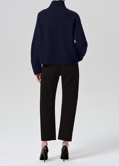A model shown from the back wearing a navy blue ribbed turtleneck sweater, black cropped trousers, and black high heels against a grey background.