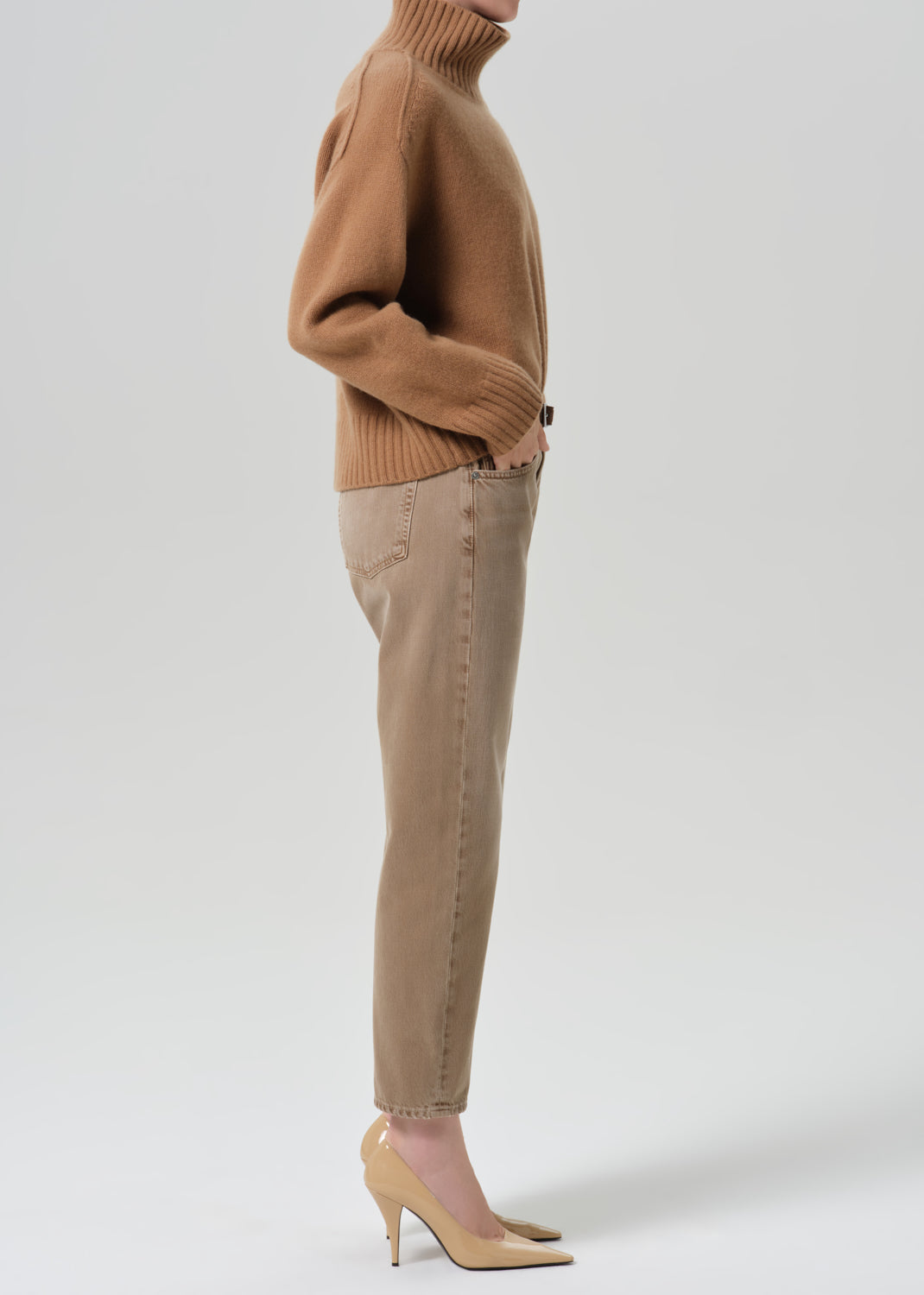 Person wearing tan straight leg pants with a brown turtleneck sweater viewed from the side on a grey background