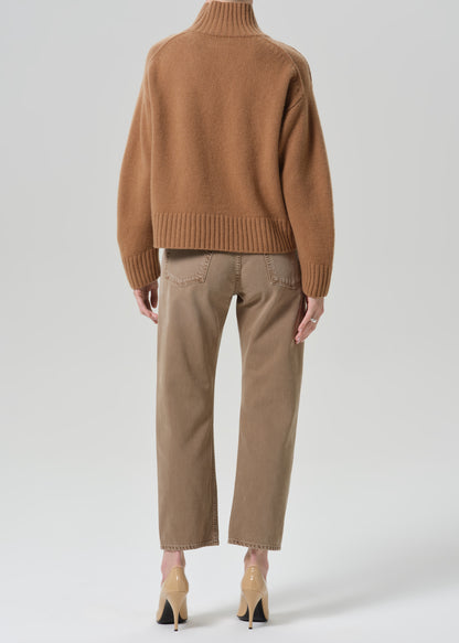 Person wearing tan straight leg pants with a brown turtleneck sweater viewed from the back on a grey background