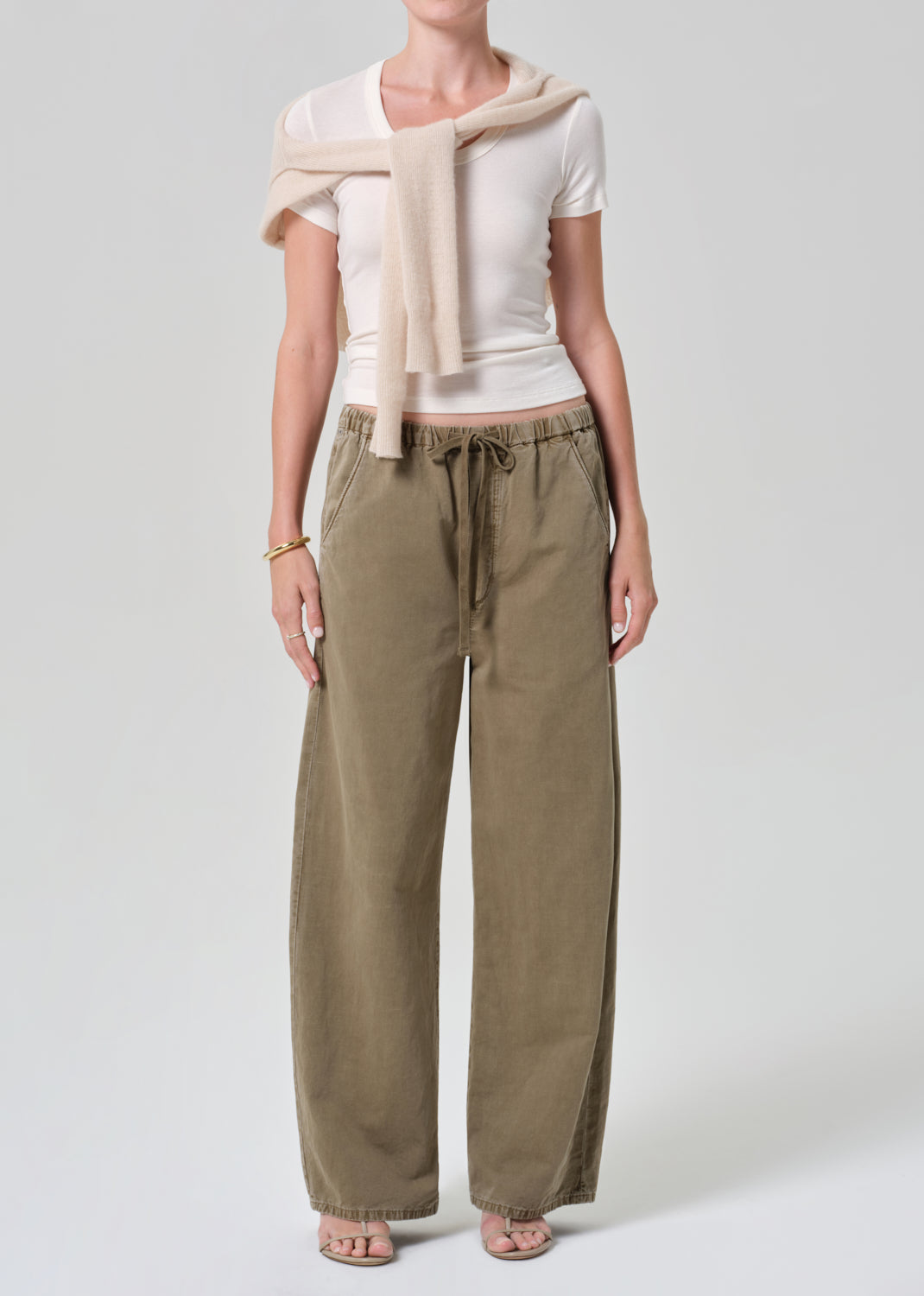Person wearing a white top and olive brown pants with a beige sweater on a light gray background