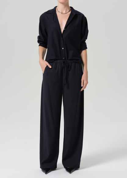 A model wearing a black wide-leg jumpsuit with a button-up shirt collar and drawstring waist, photographed against a grey background.