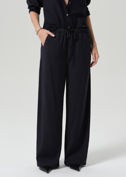 A model wearing a black wide-leg jumpsuit with a drawstring waist, paired with a black collared top, shown against a grey background.