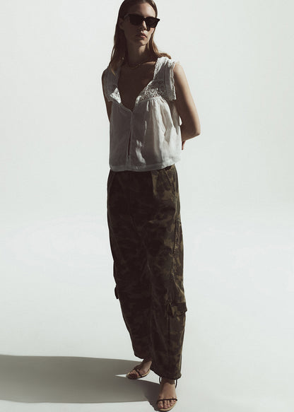 Woman wearing a white sleeveless top and camo cargo pants on a plain background