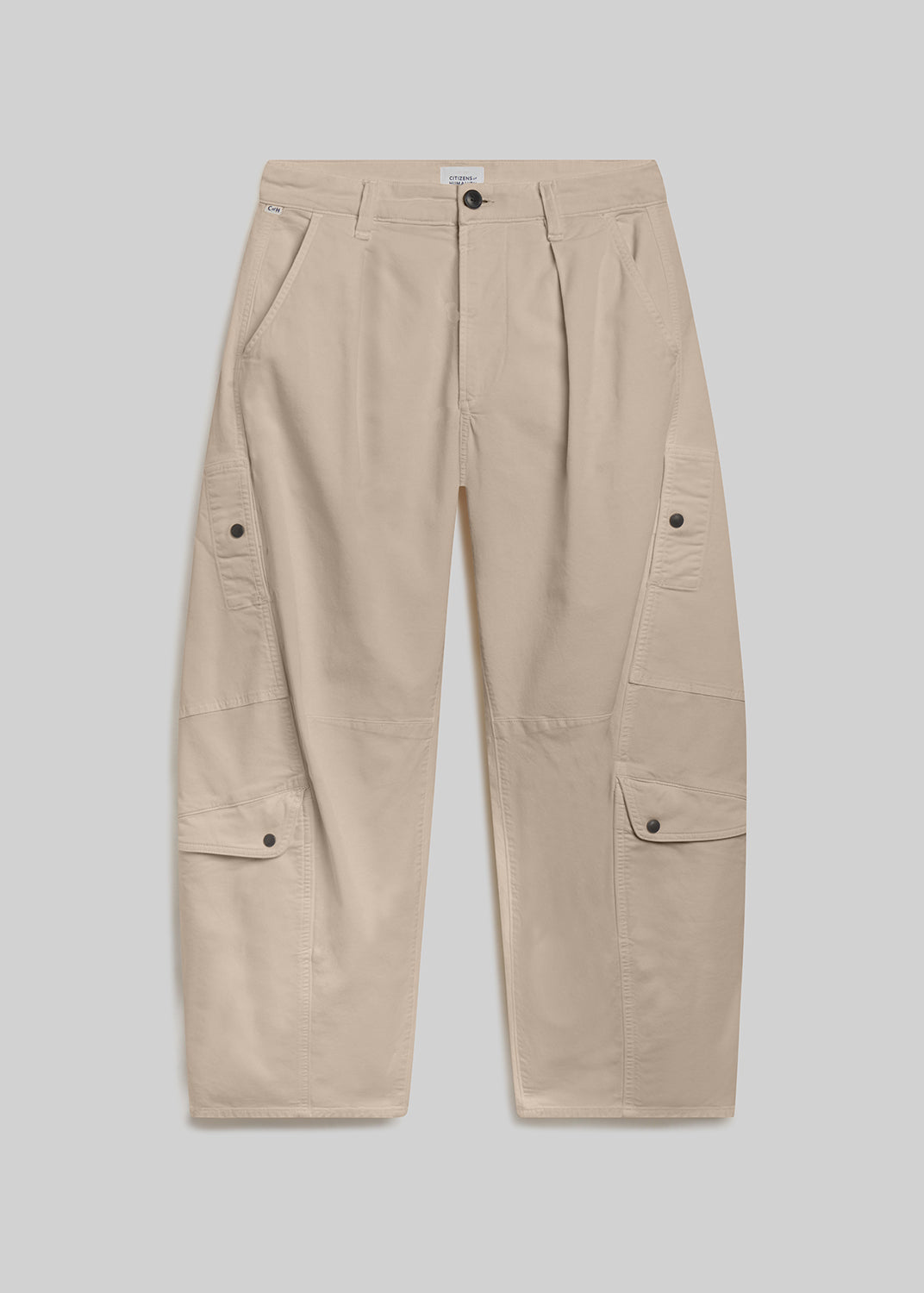 Beige utility pants with a relaxed barrel-leg silhouette, front pleats, belt loops, button closure, and oversized cargo pockets on both legs, front flat lay view on a grey background.