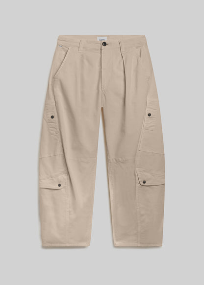 Beige utility pants with a relaxed barrel-leg silhouette, front pleats, belt loops, button closure, and oversized cargo pockets on both legs, front flat lay view on a grey background.