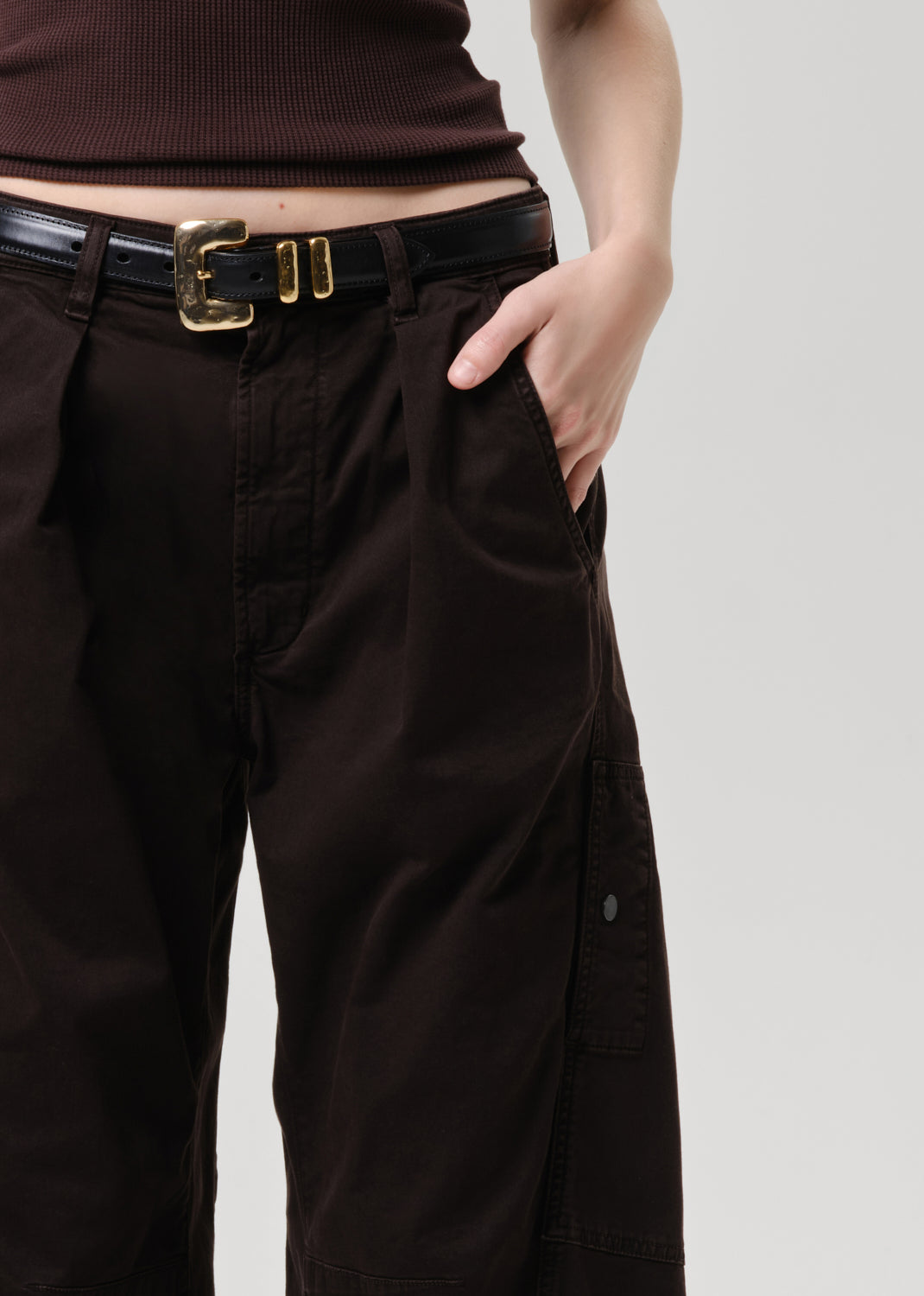 Close-up of a person wearing dark brown wide-leg cargo pants with pleats, a black leather belt with a gold buckle, and a dark brown ribbed top against a grey background.