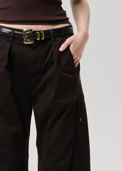 Close-up of a person wearing dark brown wide-leg cargo pants with pleats, a black leather belt with a gold buckle, and a dark brown ribbed top against a grey background.