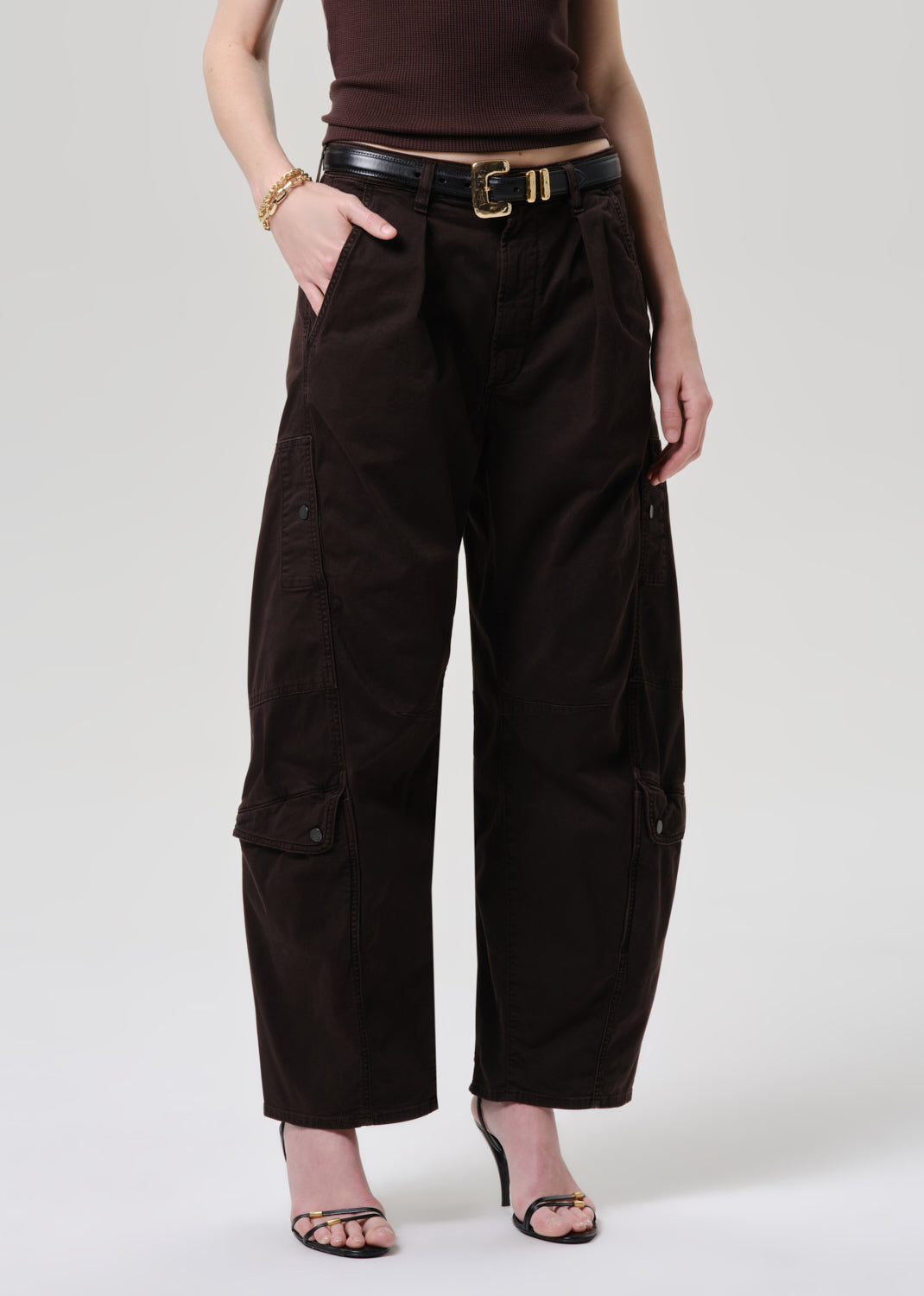 A model wearing dark brown wide-leg cargo pants with side flap pockets, styled with a black belt, a dark brown top, and black heeled sandals against a grey background.
