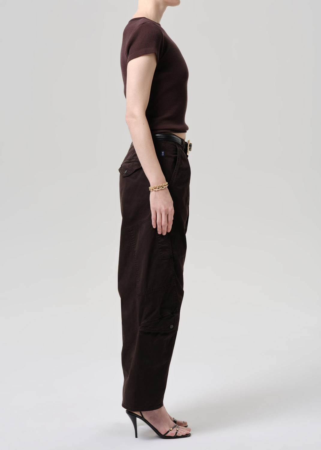 A model shown in profile wearing a dark brown short-sleeve top, dark brown wide-leg cargo pants, a black belt with a gold buckle, and black high-heeled sandals against a grey background.