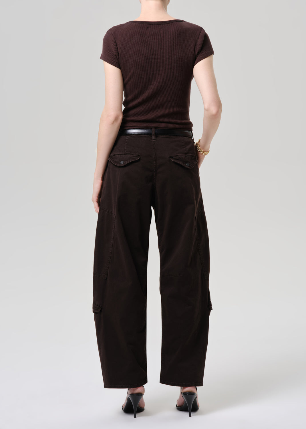 A model shown from the back wearing dark brown wide-leg cargo pants with flap pockets, a dark brown short-sleeve top, a black belt, and black heels, set against a grey background.