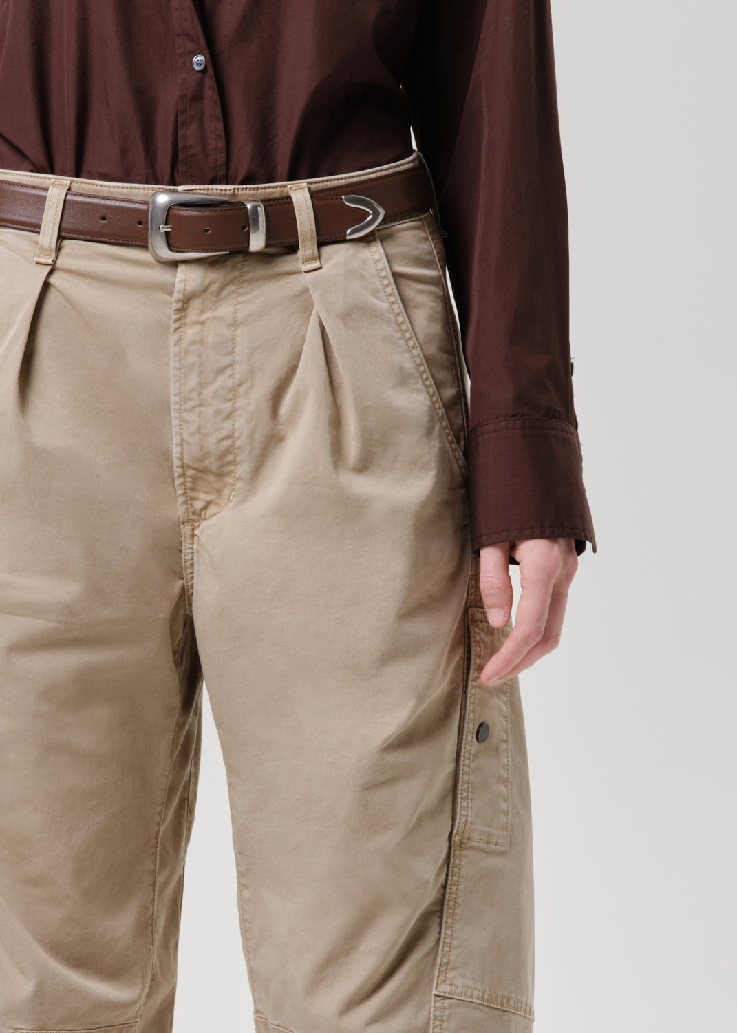 A close-up of a person wearing beige pleated cargo pants with a brown belt, paired with a dark brown button-up shirt, against a grey background.